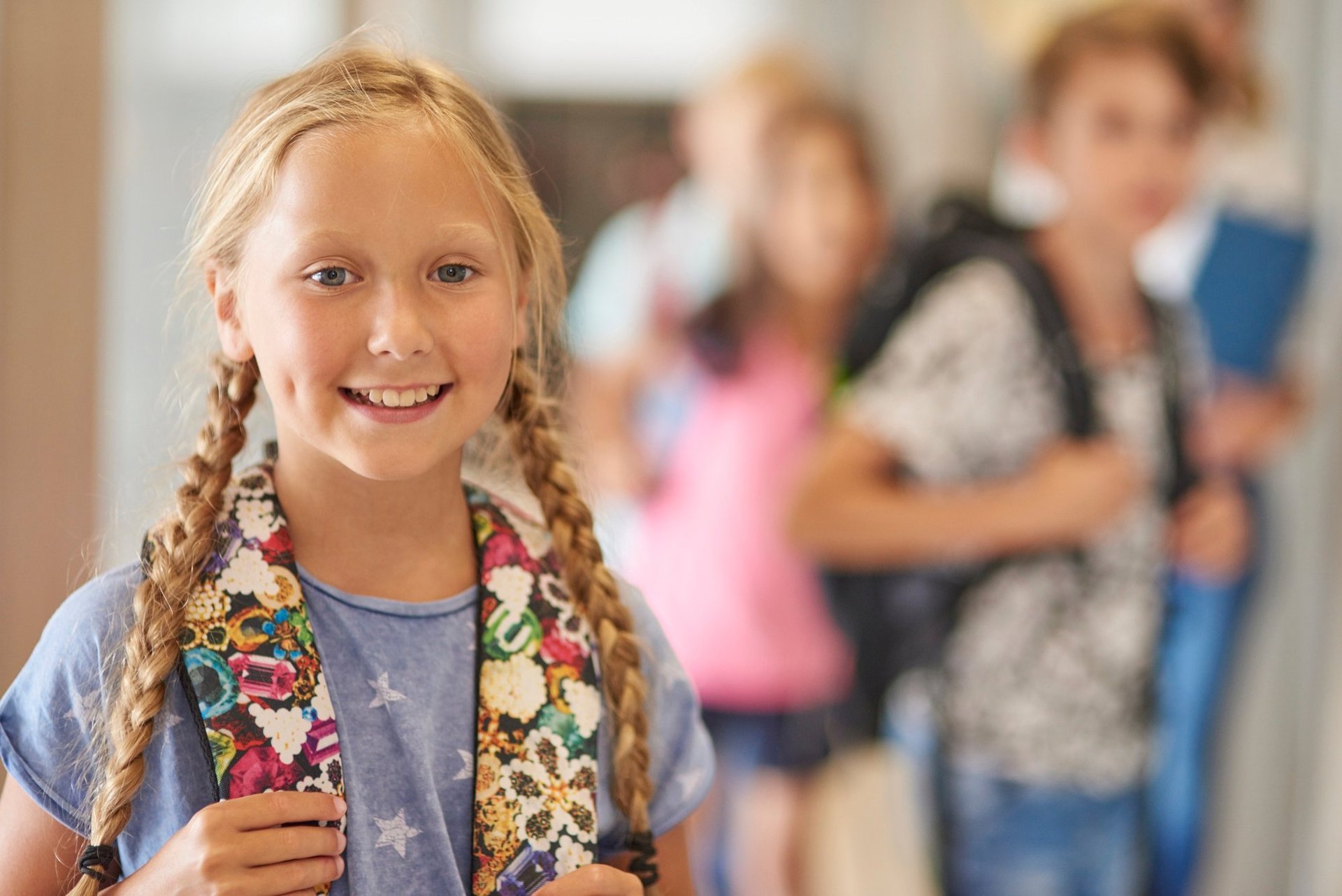 Young girl with braided blonde hair smiling at camera, wearing blue shirt and colorful scarf, with blurred students and school hallway in background